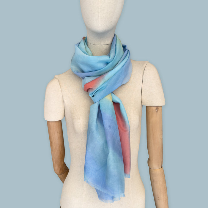 Mannequin wearing scarf on a blue background. Hand-painted silk cotton scarf by Otta-Italy, measuring 70 x 190 cm, featuring 
blue, yellow, red, orange and green shades. Luxury Italian pure silk cotton blend accessory, soft, fresh and lightweight scarf for women and men.
