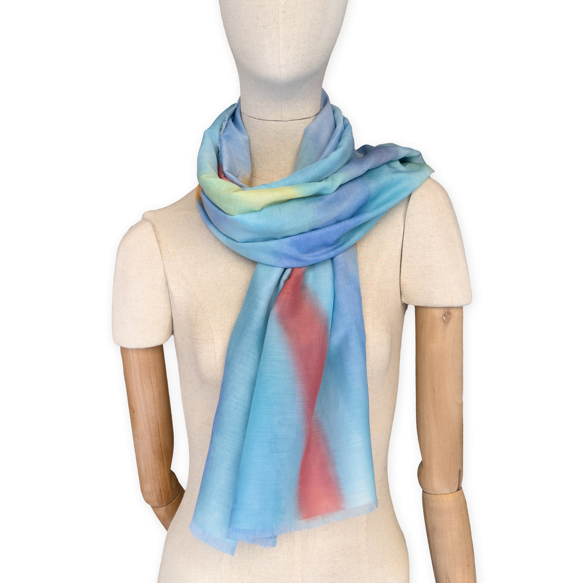 Mannequin wearing a hand-painted Otta silk cotton blend scarf on a white background in 
blue, yellow, red, orange and green tones. soft, fresh and lightweight, and elegant accessory measuring 70 x 190 cm. Unique artisan scarf with hand-fringed edges, perfect for women and men.
