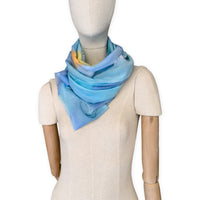 Mannequin wearing a hand-painted Otta silk cotton blend scarf on a white background in 
blue, yellow, red, orange and green tones. soft, fresh and lightweight, and elegant accessory measuring 70 x 190 cm. Unique artisan scarf with hand-fringed edges, perfect for women and men.
