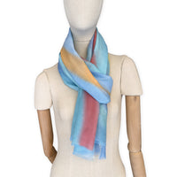 Mannequin wearing a hand-painted Otta silk cotton blend scarf on a white background in 
blue, yellow, red, orange and green tones. soft, fresh and lightweight, and elegant accessory measuring 70 x 190 cm. Unique artisan scarf with hand-fringed edges, perfect for women and men.
