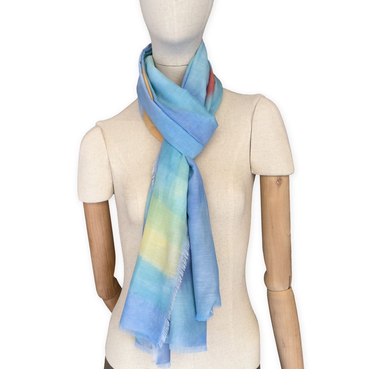 Mannequin wearing a hand-painted Otta silk cotton blend scarf on a white background in 
blue, yellow, red, orange and green tones. soft, fresh and lightweight, and elegant accessory measuring 70 x 190 cm. Unique artisan scarf with hand-fringed edges, perfect for women and men.
