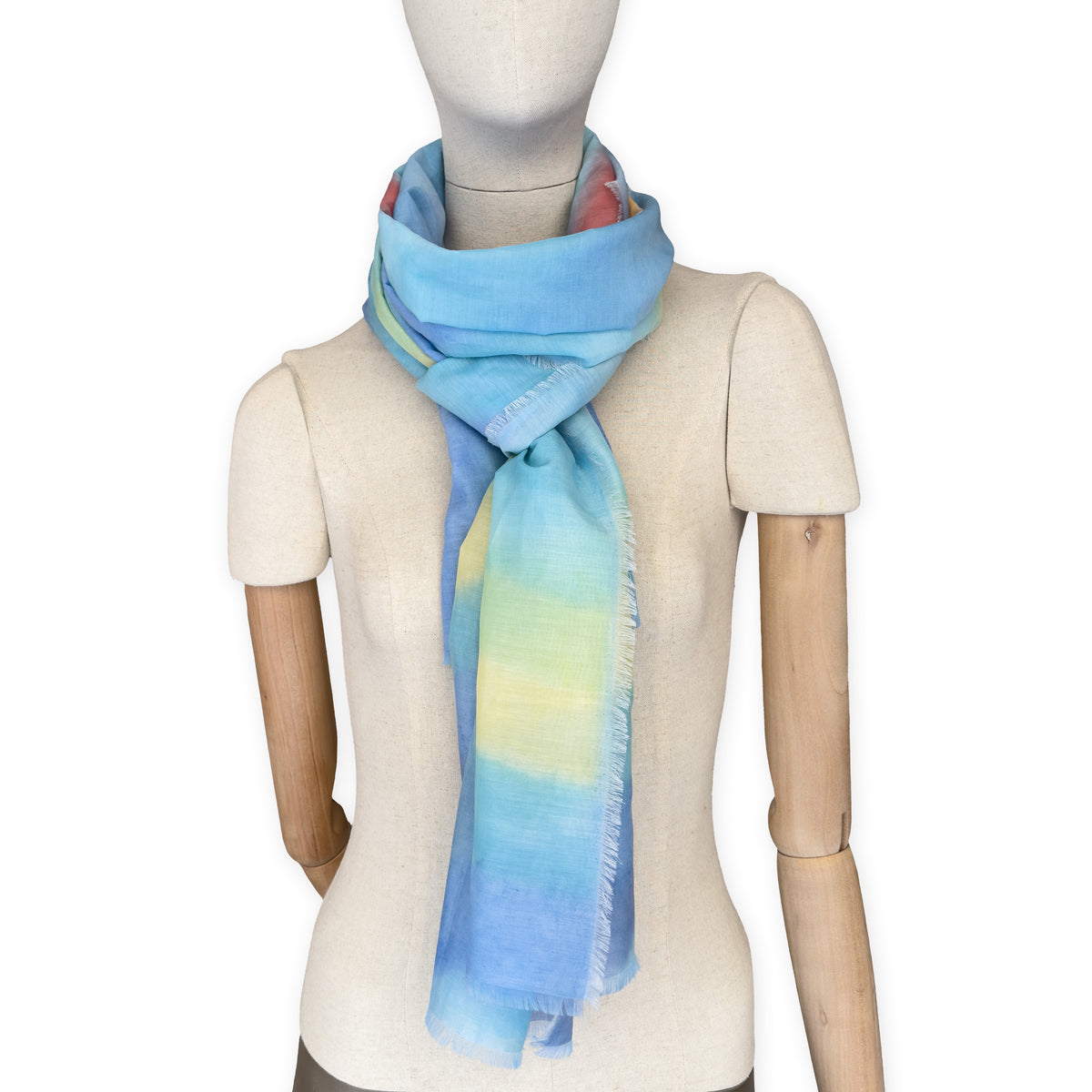 Mannequin wearing a hand-painted Otta silk cotton blend scarf on a white background in 
blue, yellow, red, orange and green tones. soft, fresh and lightweight, and elegant accessory measuring 70 x 190 cm. Unique artisan scarf with hand-fringed edges, perfect for women and men.
