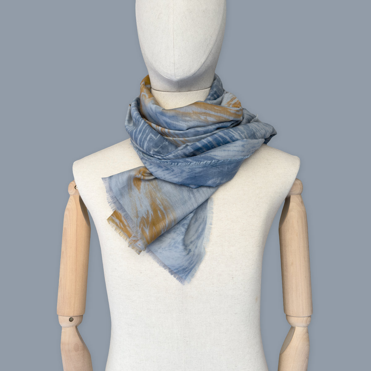 Mannequin wearing scarf on a blue background. Hand-painted silk cotton scarf by Otta-Italy, measuring 67 x 193 cm, featuring 
blue, gray and orange shades. Luxury Italian pure silk cotton blend accessory, soft, fresh and lightweight scarf for women and men.
