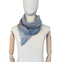 Mannequin wearing a hand-painted Otta silk cotton blend scarf on a white background in 
blue, gray and orange e tones. soft, fresh and lightweight, and elegant accessory measuring 67 x 193 cm. Unique artisan scarf with hand-fringed edges, perfect for women and men.
