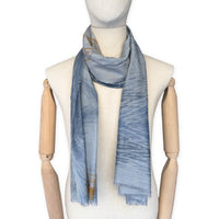 Mannequin wearing a hand-painted Otta silk cotton blend scarf on a white background in 
blue, gray and orange e tones. soft, fresh and lightweight, and elegant accessory measuring 67 x 193 cm. Unique artisan scarf with hand-fringed edges, perfect for women and men.
