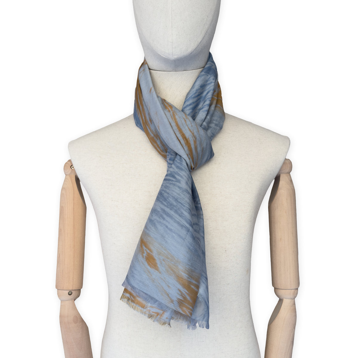 Mannequin wearing a hand-painted Otta silk cotton blend scarf on a white background in 
blue, gray and orange e tones. soft, fresh and lightweight, and elegant accessory measuring 67 x 193 cm. Unique artisan scarf with hand-fringed edges, perfect for women and men.
