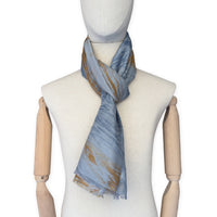 Mannequin wearing a hand-painted Otta silk cotton blend scarf on a white background in 
blue, gray and orange e tones. soft, fresh and lightweight, and elegant accessory measuring 67 x 193 cm. Unique artisan scarf with hand-fringed edges, perfect for women and men.
