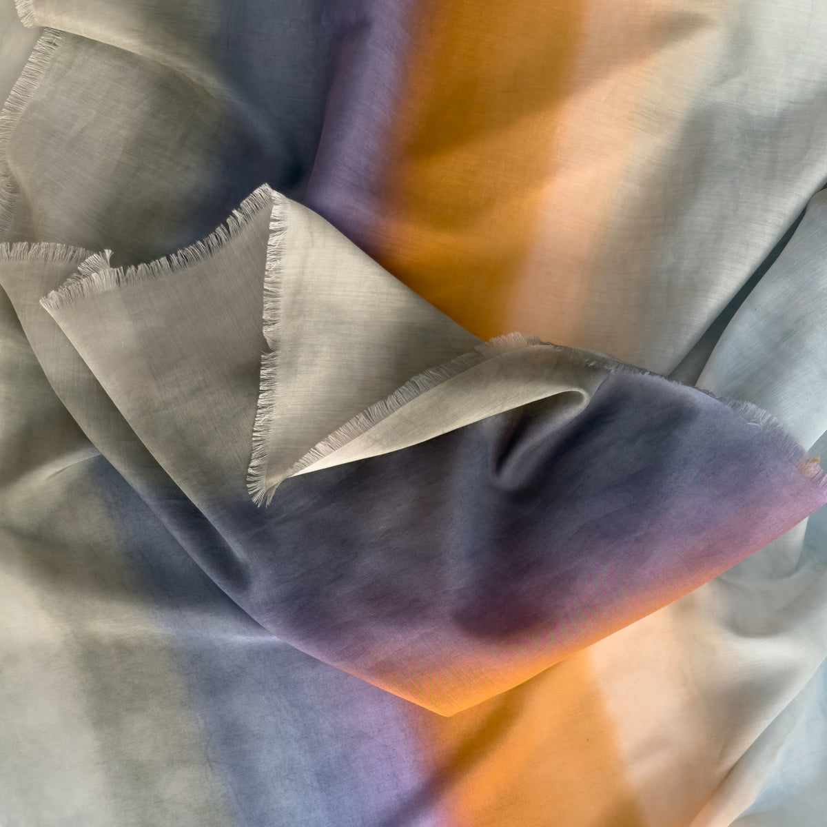 Close-up of a hand-painted Otta silk cotton blend scarf draped softly, showcasing gentle shades of 
gray, purple and orange. Lightweight artisan accessory with delicate hand-fringed edges, measuring 70 x 190 cm.
