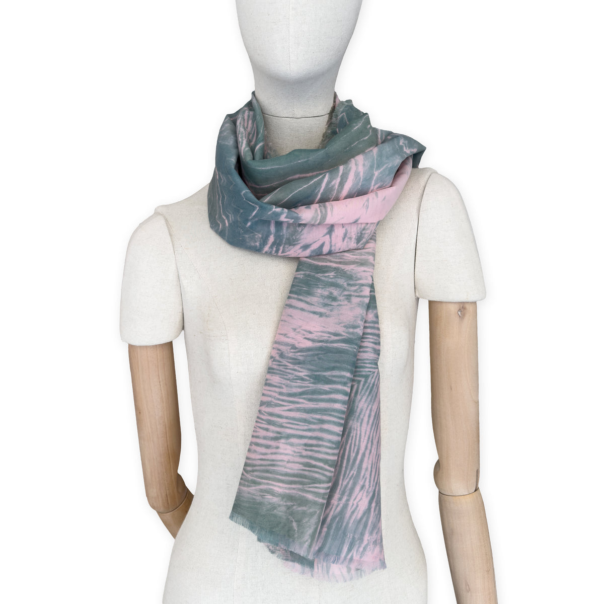 Mannequin wearing a hand-painted Otta silk cotton blend scarf on a white background in 
pink and green e tones. soft, fresh and lightweight, and elegant accessory measuring 70 x 190 cm. Unique artisan scarf with hand-fringed edges, perfect for women and men.
