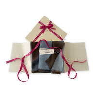 Luxury gift box with a pink ribbon containing a hand-painted Otta pure cashmere scarf on a white background.
