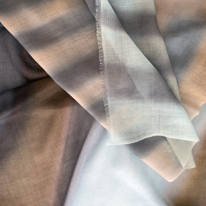 Close-up of hand-painted Otta pure cashmere scarf showcasing soft light blue, beige, brown and gray shades with delicate hand-fringed edges.
