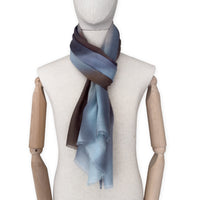 Mannequin wearing an elegant hand-painted Otta pure cashmere scarf on a white background. Warm blue, beige, orange, gray and brown tones.
