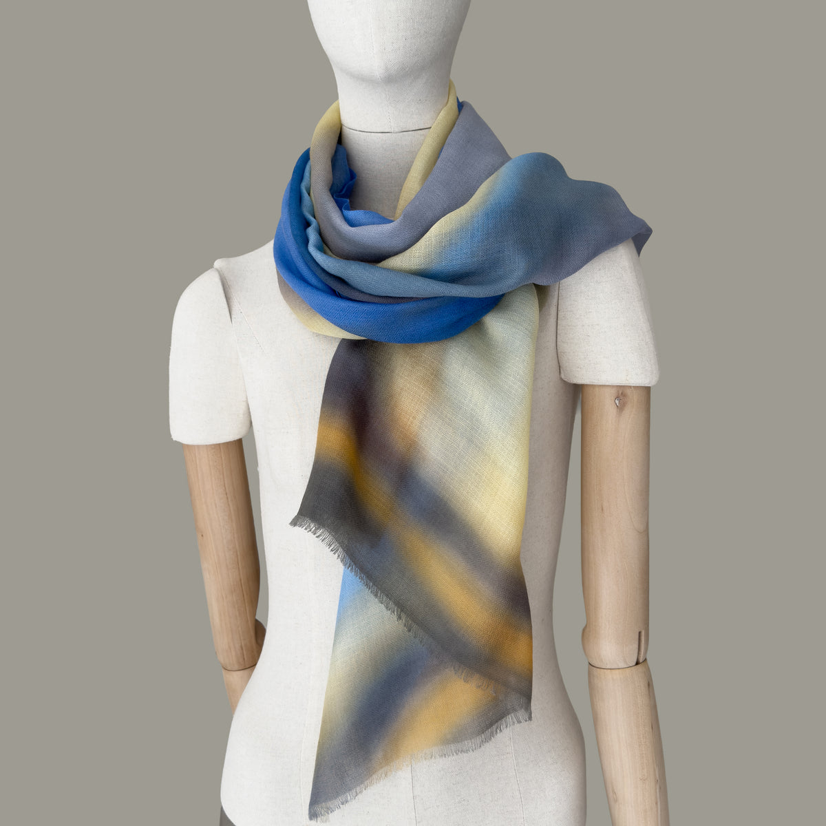 Mannequin wearing a hand-painted pure cashmere scarf by Otta-Italy, gray background. Blue, yellow, orange, gray and brown shades. 67x195 cm.
