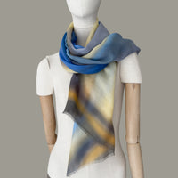 Mannequin wearing a hand-painted pure cashmere scarf by Otta-Italy, gray background. Blue, yellow, orange, gray and brown shades. 67x195 cm.
