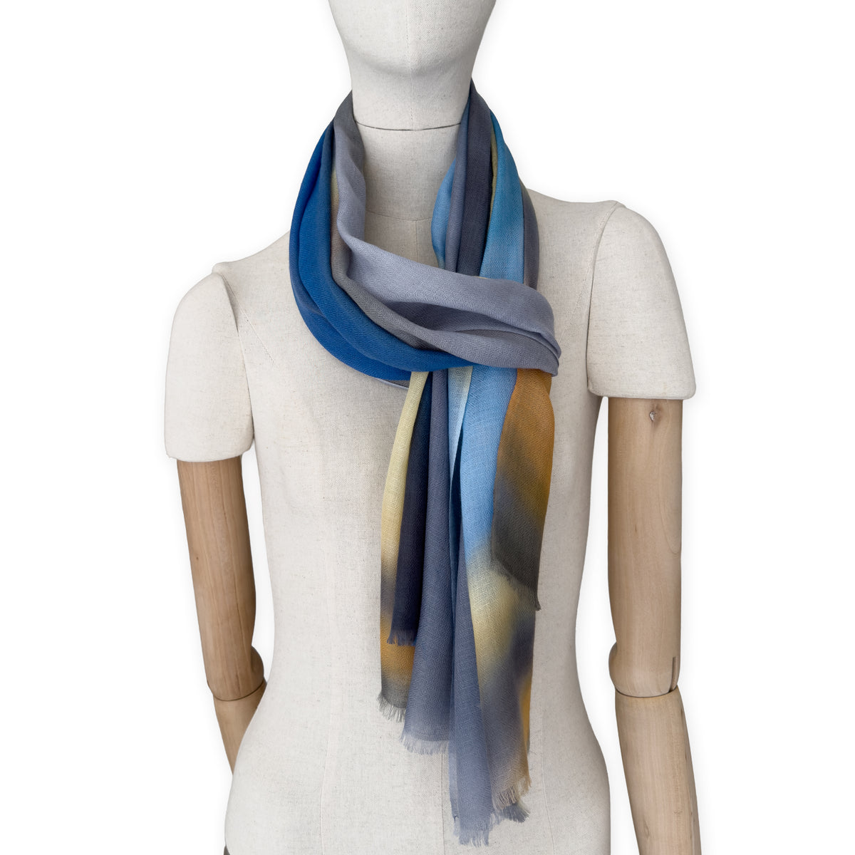 Mannequin wearing an elegant hand-painted Otta pure cashmere scarf on a white background. Warm blue, yellow, orange, gray and brown tones.
