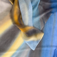 Close-up of hand-painted Otta pure cashmere scarf showcasing soft blue, yellow, orange, gray and brown shades with delicate hand-fringed edges.
