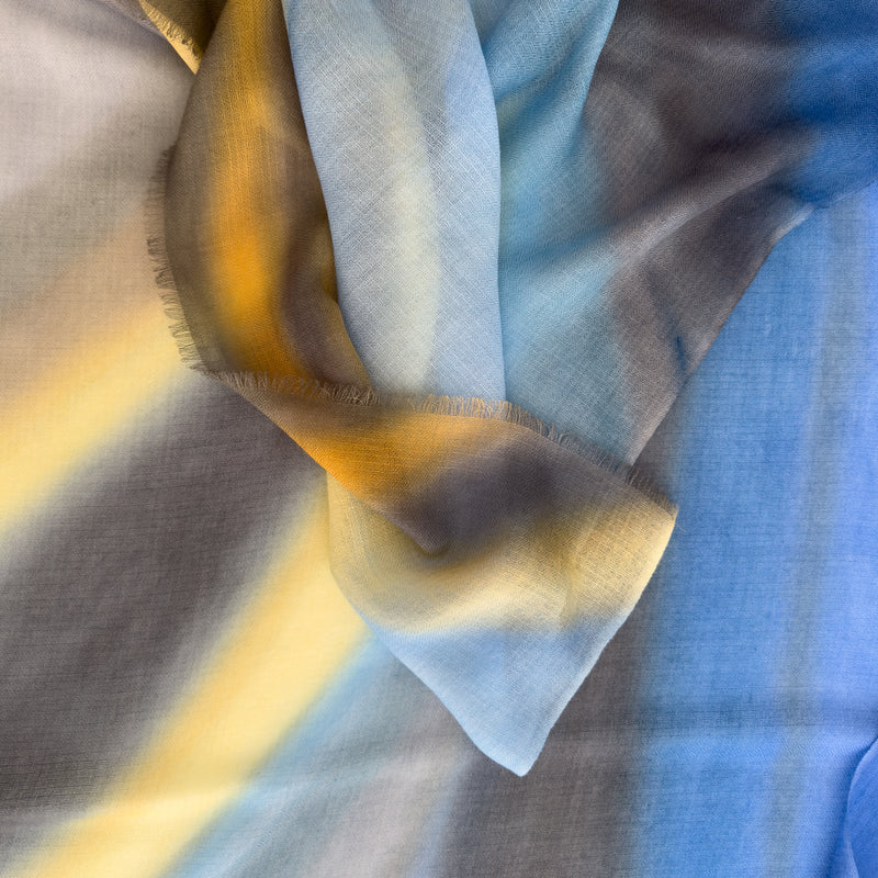 Close-up of hand-painted Otta pure cashmere scarf showcasing soft blue, yellow, orange, gray and brown shades with delicate hand-fringed edges.
