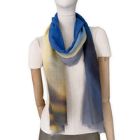 Mannequin wearing an elegant hand-painted Otta pure cashmere scarf on a white background. Warm blue, yellow, orange, gray and brown tones.
