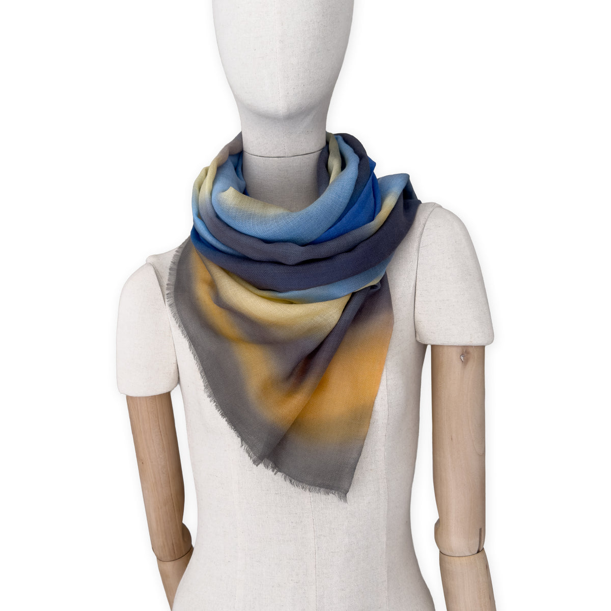 Mannequin wearing an elegant hand-painted Otta pure cashmere scarf on a white background. Warm blue, yellow, orange, gray and brown tones.
