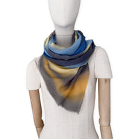 Mannequin wearing an elegant hand-painted Otta pure cashmere scarf on a white background. Warm blue, yellow, orange, gray and brown tones.
