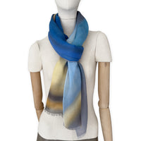 Mannequin wearing an elegant hand-painted Otta pure cashmere scarf on a white background. Warm blue, yellow, orange, gray and brown tones.
