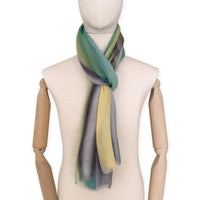 Mannequin wearing a hand-painted Otta cashmere scarf on a white background in 
green, yellow, gray and brown tones. Lightweight, warm, and elegant accessory measuring 68 x 202 cm. Unique artisan scarf with hand-fringed edges, perfect for women and men.
