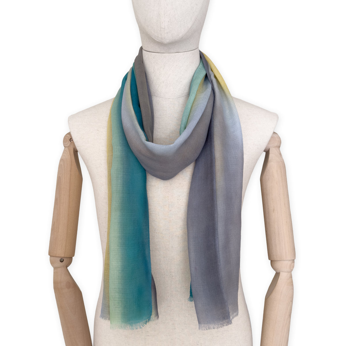 Mannequin wearing a hand-painted Otta cashmere scarf on a white background in 
green, yellow, gray and brown tones. Lightweight, warm, and elegant accessory measuring 68 x 202 cm. Unique artisan scarf with hand-fringed edges, perfect for women and men.
