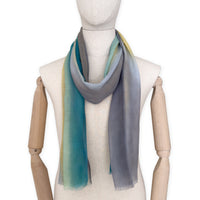 Mannequin wearing a hand-painted Otta cashmere scarf on a white background in 
green, yellow, gray and brown tones. Lightweight, warm, and elegant accessory measuring 68 x 202 cm. Unique artisan scarf with hand-fringed edges, perfect for women and men.

