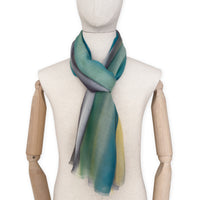 Mannequin wearing a hand-painted Otta cashmere scarf on a white background in 
green, yellow, gray and brown tones. Lightweight, warm, and elegant accessory measuring 68 x 202 cm. Unique artisan scarf with hand-fringed edges, perfect for women and men.
