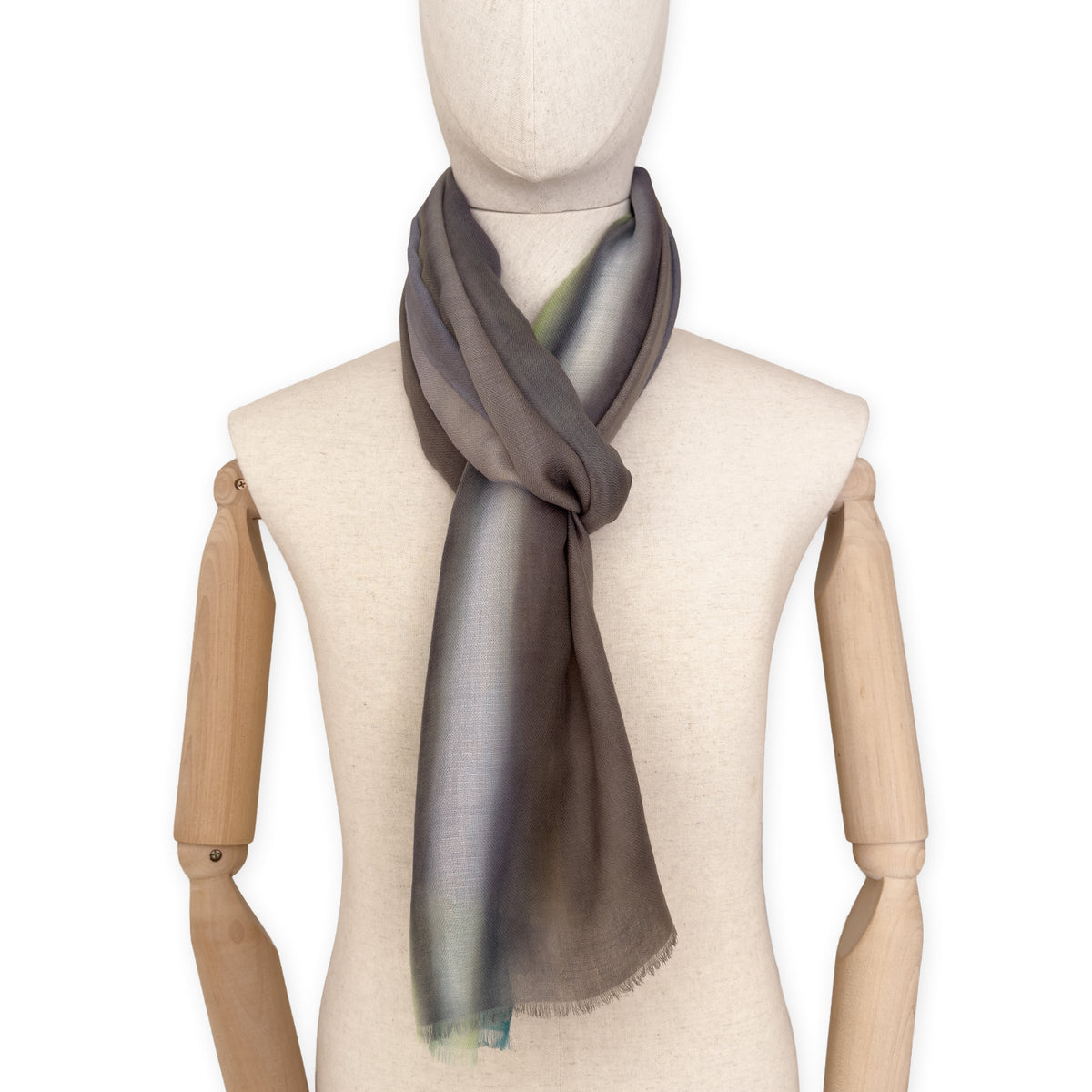 Mannequin wearing a hand-painted Otta cashmere scarf on a white background in 
green, yellow, gray and brown tones. Lightweight, warm, and elegant accessory measuring 68 x 202 cm. Unique artisan scarf with hand-fringed edges, perfect for women and men.
