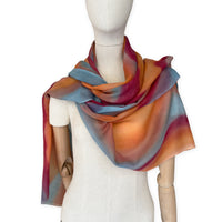 Mannequin wearing a hand-painted Otta cashmere scarf on a white background in 
orange, red and blue tones. Lightweight, warm, and elegant accessory measuring 69 x 201 cm. Unique artisan scarf with hand-fringed edges, perfect for women and men.
