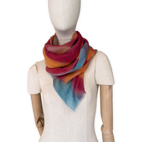 Mannequin wearing a hand-painted Otta cashmere scarf on a white background in 
orange, red and blue tones. Lightweight, warm, and elegant accessory measuring 69 x 201 cm. Unique artisan scarf with hand-fringed edges, perfect for women and men.
