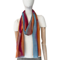 Mannequin wearing a hand-painted Otta cashmere scarf on a white background in 
orange, red and blue tones. Lightweight, warm, and elegant accessory measuring 69 x 201 cm. Unique artisan scarf with hand-fringed edges, perfect for women and men.
