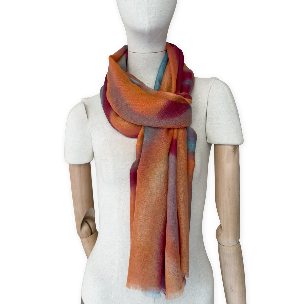 Mannequin wearing a hand-painted Otta cashmere scarf on a white background in 
orange, red and blue tones. Lightweight, warm, and elegant accessory measuring 69 x 201 cm. Unique artisan scarf with hand-fringed edges, perfect for women and men.
