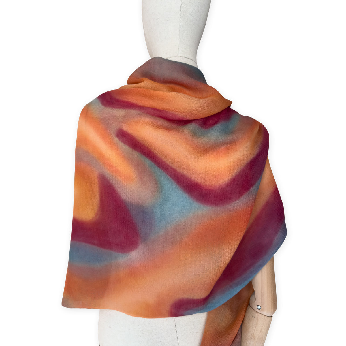 Mannequin wearing a hand-painted Otta cashmere scarf on a white background in 
orange, red and blue tones. Lightweight, warm, and elegant accessory measuring 69 x 201 cm. Unique artisan scarf with hand-fringed edges, perfect for women and men.
