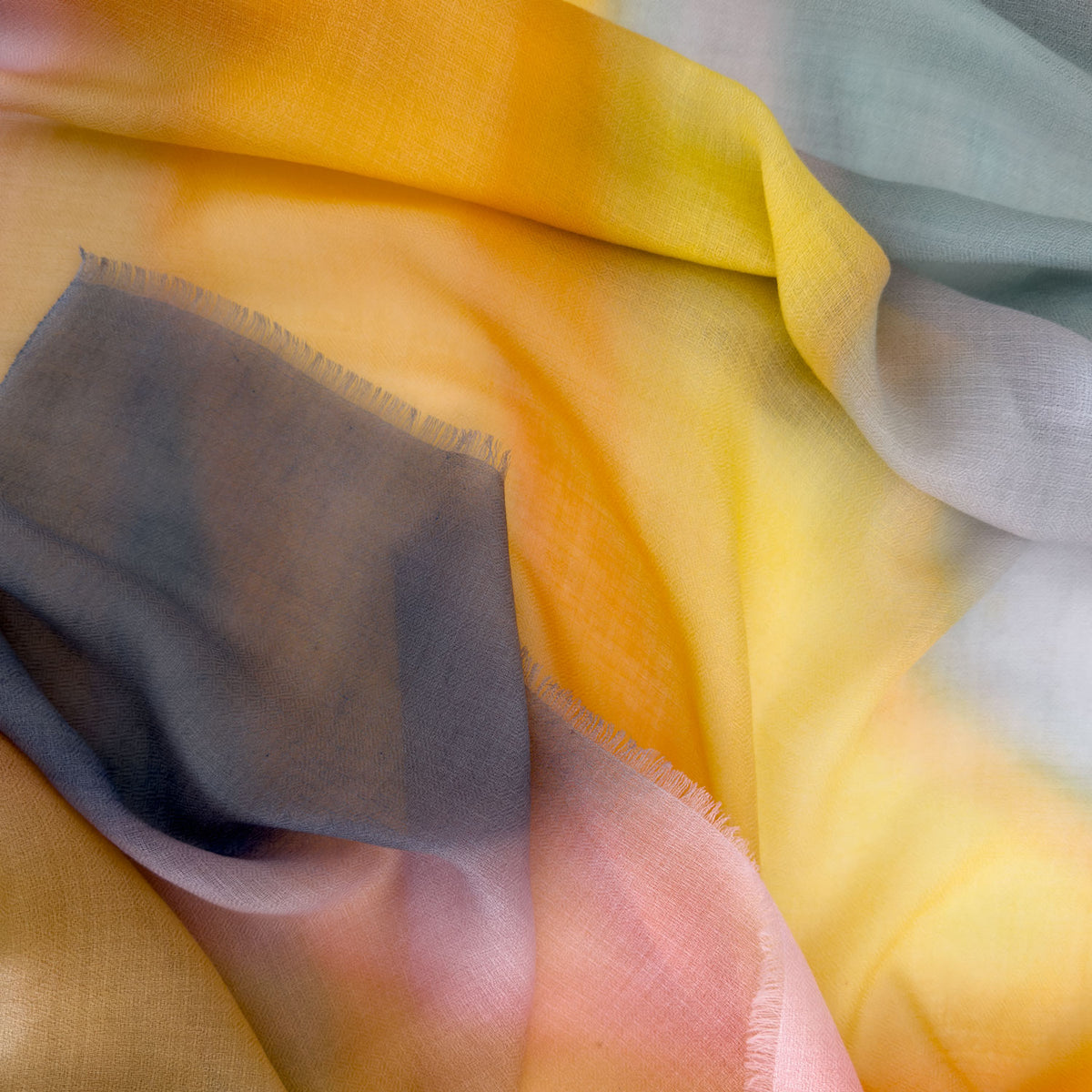 Close-up of hand-painted Otta pure cashmere scarf showcasing soft yellow, orange, pink and gray shades with delicate hand-fringed edges.
