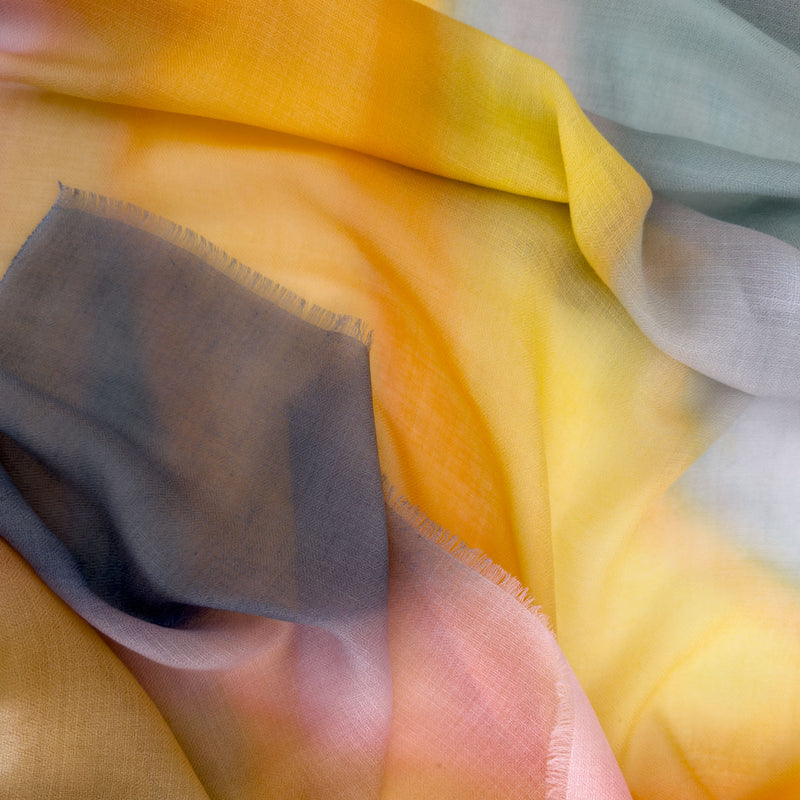 Close-up of hand-painted Otta pure cashmere scarf showcasing soft yellow, orange, pink and gray shades with delicate hand-fringed edges.

