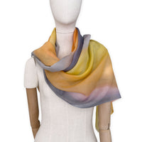 Mannequin wearing an elegant hand-painted Otta pure cashmere scarf on a white background. Warm yellow, orange, pink and gray tones.
