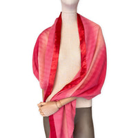 Mannequin wearing a hand-painted Otta cashmere & wool blend scarf on a white background in 
red, fuchsia and pink tones. Lightweight, warm, and elegant accessory measuring 57 x 180 cm. Unique artisan scarf with hand-fringed edges, perfect for women and men.
