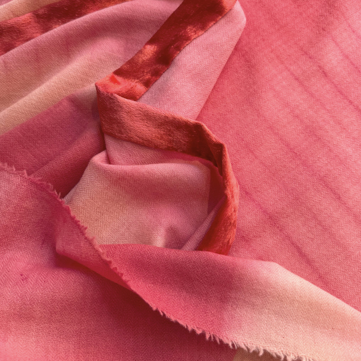 Close-up of a hand-painted Otta cashmere & wool scarf draped softly, showcasing gentle shades of 
red, fuchsia and pink. Lightweight artisan accessory with delicate hand-fringed edges, measuring 57 x 180 cm.
