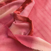 Close-up of a hand-painted Otta cashmere & wool scarf draped softly, showcasing gentle shades of 
red, fuchsia and pink. Lightweight artisan accessory with delicate hand-fringed edges, measuring 57 x 180 cm.
