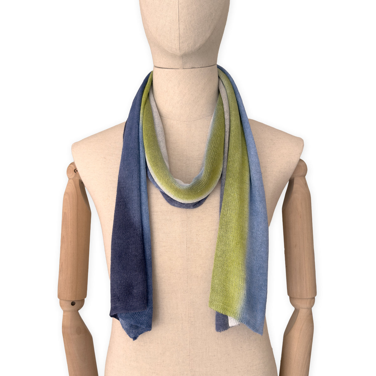 Mannequin wearing a hand-painted Otta pure linen scarf on a white background in 
blue, midnight blue, indigo, light blue, green tones. soft, fresh and lightweight, and elegant accessory measuring 34 x 192 cm. Unique artisan scarf with hand-sewn edges, perfect for women and men.
