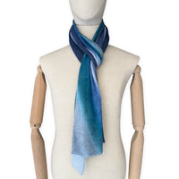 Mannequin wearing a colorful and lightweight linen scarf on a white background. Linen scarf is in shades of blue, midnight blue, light blue, turquoise and indigo colors. It is hand-painted scarf in measures 32 x 200 cm by otta-italy scarves brand. Luxury Italian linen, soft and lightweight scarf for women and men.