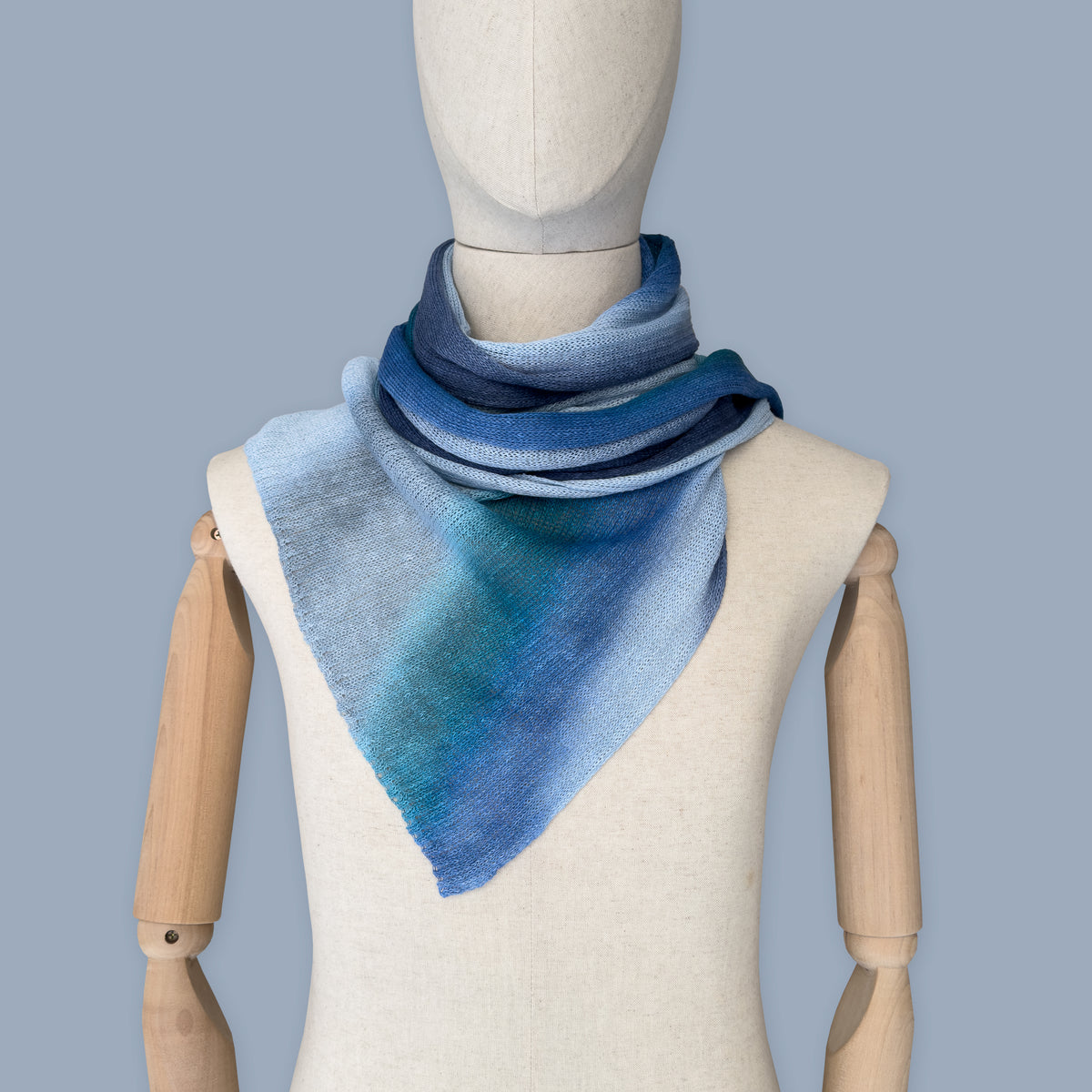 Mannequin wearing a colorful and lightweight linen scarf on a gray background. Linen scarf is in shades of blue, midnight blue, light blue, turquoise and indigo colors. It is hand-painted scarf in measures 32 x 200 cm by otta-italy scarves brand. Luxury Italian linen, soft and lightweight scarf for women and men.