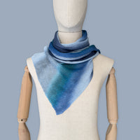 Mannequin wearing a colorful and lightweight linen scarf on a gray background. Linen scarf is in shades of blue, midnight blue, light blue, turquoise and indigo colors. It is hand-painted scarf in measures 32 x 200 cm by otta-italy scarves brand. Luxury Italian linen, soft and lightweight scarf for women and men.