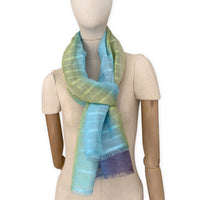 Mannequin wearing a hand-painted Otta pure linen scarf on a white background in 
blue, turquoise, green and purple tones. soft, fresh and lightweight, and elegant accessory measuring 70 x 190 cm. Unique artisan scarf with hand-sewn edges, perfect for women and men.

