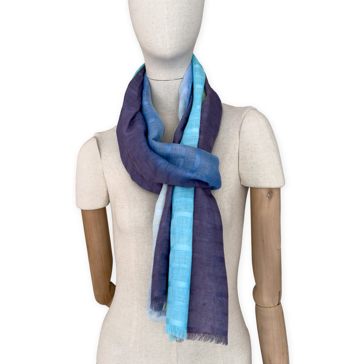 Mannequin wearing a hand-painted Otta pure linen scarf on a white background in 
blue, turquoise, green and purple tones. soft, fresh and lightweight, and elegant accessory measuring 70 x 190 cm. Unique artisan scarf with hand-sewn edges, perfect for women and men.
