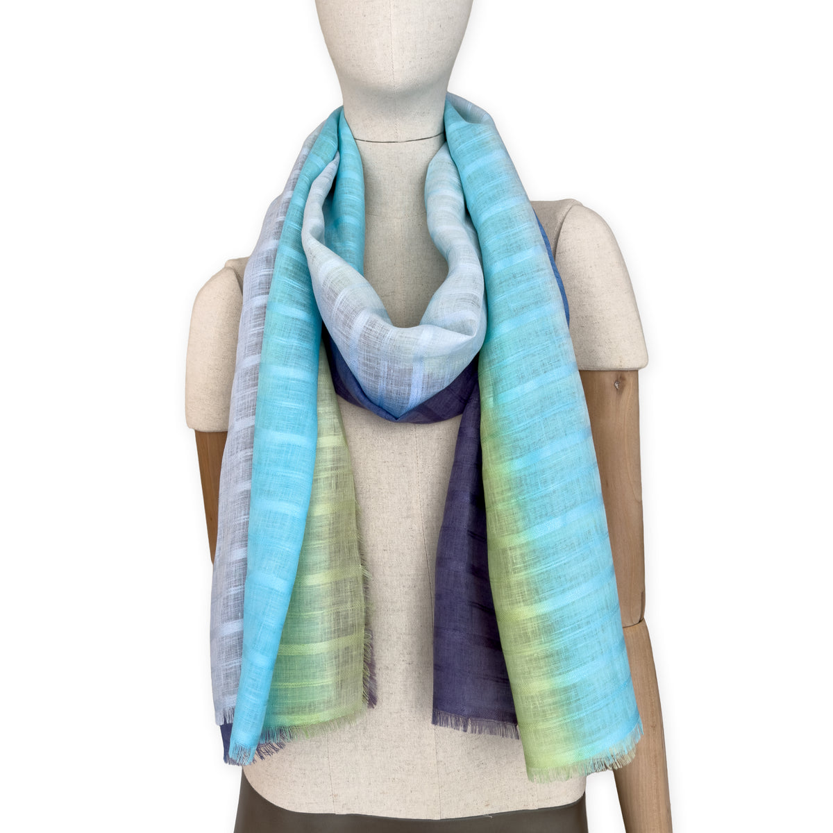 Mannequin wearing a hand-painted Otta pure linen scarf on a white background in 
blue, turquoise, green and purple tones. soft, fresh and lightweight, and elegant accessory measuring 70 x 190 cm. Unique artisan scarf with hand-sewn edges, perfect for women and men.
