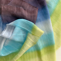 Close-up of soft fabric, a hand-painted Otta pure linen scarf draped softly, showcasing gentle shades of 
blue, turquoise, green and purple. Lightweight artisan accessory with delicate hand-sewn edges, measuring 70 x 190 cm.
