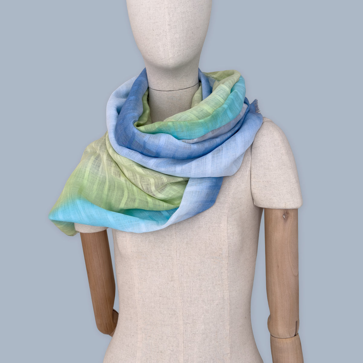 
Mannequin wearing scarf on a pastel blue background. Hand-painted linen scarf by Otta-Italy, 70 x 190 cm, featuring 
blue, turquoise, green and purple. Luxury Italian pure linen accessory, soft, fresh and lightweight scarf for women and men.
